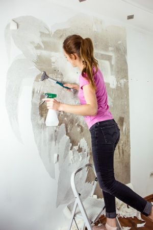 Junge Frau entfernt die Tapete mit Tapetenablöser aus Sprühflasche und Spachtel